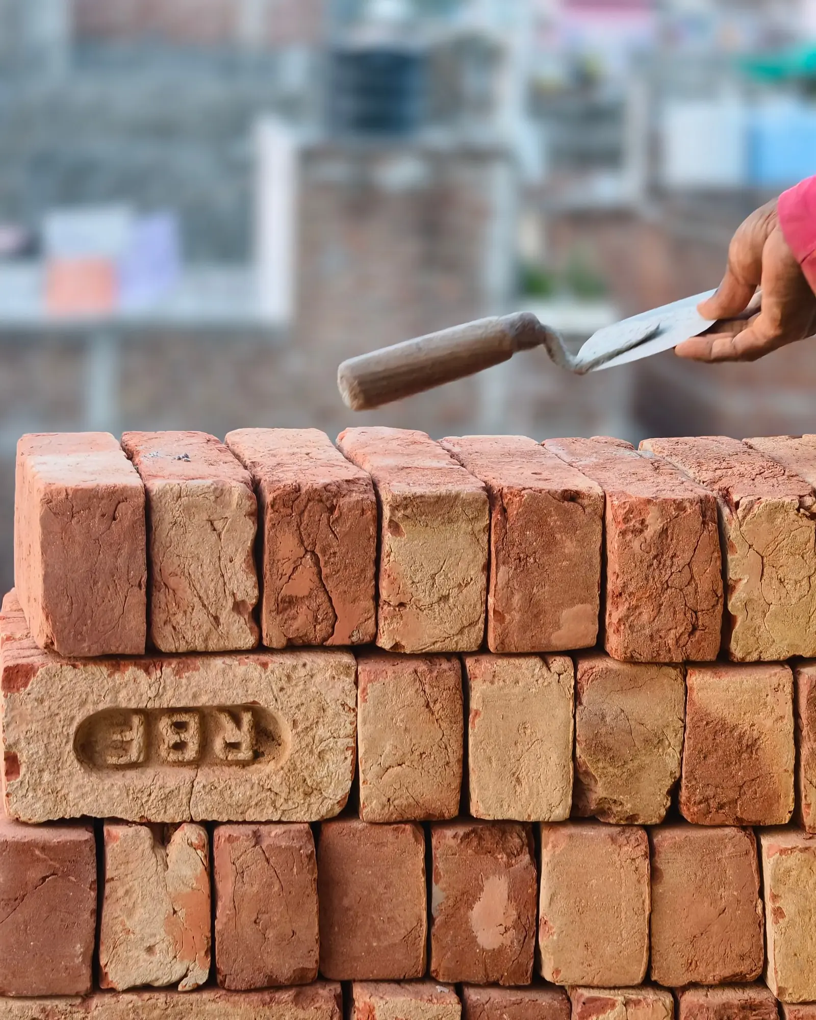 Gloved hand holding a mason's trowel over a stack of red bricks
